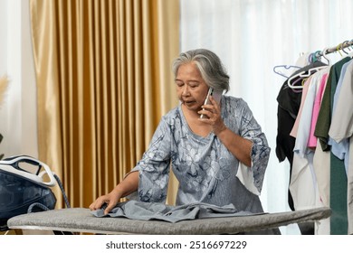 Senior Woman woman multitasks by ironing clothes while talking with her smartphone calling, surrounded by hanging garments in a bright room. - Powered by Shutterstock