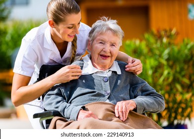 Senior Woman In Nursing Home With Nurse In Garden Sitting In Wheelchair 