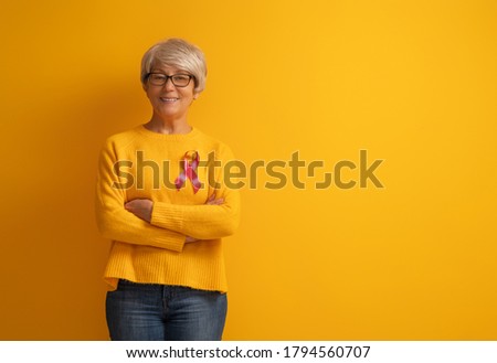 Senior woman on color yellow background. Pink ribbon like a symbol of breast cancer awareness. Support people living with tumor illness.