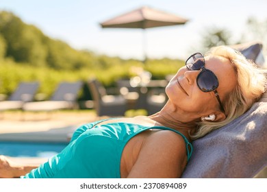 Senior Woman On Holiday In Swimming Costume Wearing Wireless Earbuds By Hotel Swimming Pool - Powered by Shutterstock