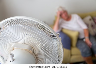 Senior woman on a sofa suffering from excessive heat during a heatwave and being cooled by an oscillating electrical fan in the foreground - Shutterstock ID 2179726515