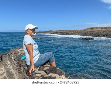 Senior woman in outdoors vacation at sea sitting on a rocky beach admiring waves splashing on the beach in a sunny windy day in Tenerife, canary island. Travel, freedom nature concepts - Powered by Shutterstock