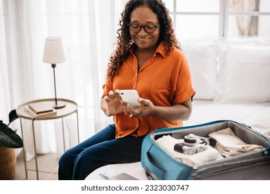 Senior woman packing her luggage with all of the essentials for her upcoming trip, using the checklist on her phone to double-check her carry-on. Retired traveler embarking on a sightseeing journey.