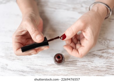 Senior woman painting her nails with dark red polish on a table.