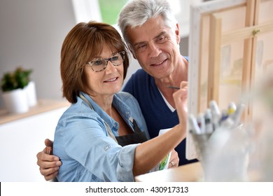 Senior Woman Painting On Canvas, Husband Beside