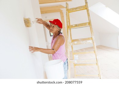 Senior Woman Painting Wall In New House. She Is Using Ladder.