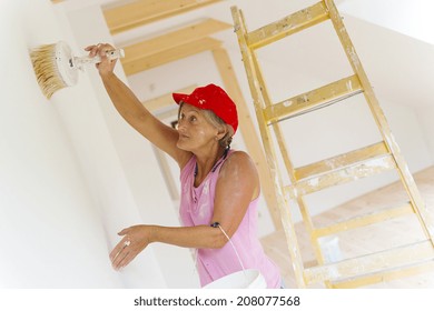 Senior Woman Painting Wall In New House. She Is Using Ladder.