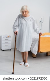 Senior Woman In Patient Gown Holding Walking Cane Near Bed