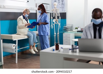 Senior Woman Patient With Protective Face Mask Signing Medical Documents During Medical Appointment In Hospital Office. Therapist Nurse Explaining Sickness Diagnosis Discussing Medication Treatment