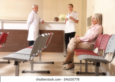 Senior Woman Patient Seated In The Hospital Waiting Room With Medical Personnel At The Desk.