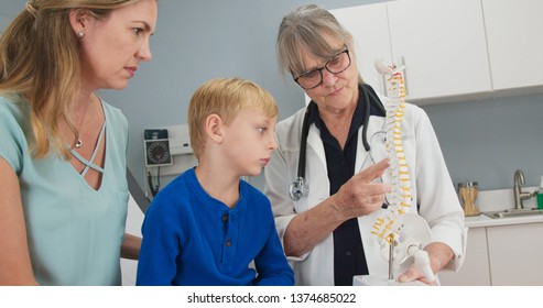 Senior Woman Pediatrician With Spine Model Talking To Little Boy Patient And His Mother About Scoliosis. Child Looking At Plastic Spinal Model With His Doctor