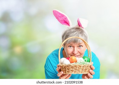 Senior Woman Peeking Out From Behind The Basket With Easter Eggs On The Natural Background