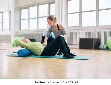 Senior woman performing back exercise on a foam roller being assisted by her personal trainer at gym. Physical therapist helping elder woman at rehab.