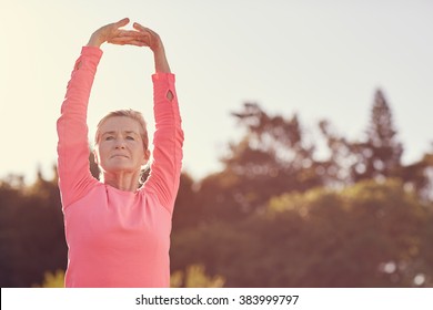 Senior Woman In A Pink Sporty Long-sleeved Top Stretching Her Arms Above Her Head To Warm-up For Some Early Morning Exercise With Gentle Sunflare Behind Her