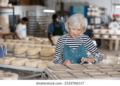 Senior woman potter in apron processing new crafted plates with tool. - Powered by Shutterstock