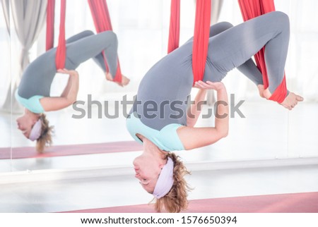 Senior woman practicing aero fly yoga in white studio with red hammocks. Concept stretching, meditation, relaxation.