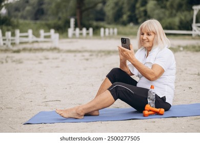 Senior woman practicing fitness on beach, using smartphone for online workout. Concept of active lifestyle, technology, and wellness in nature. - Powered by Shutterstock