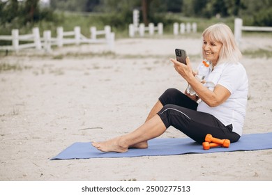 Senior woman practicing fitness on beach, using smartphone for online workout. Concept of active lifestyle, technology, and wellness in nature. - Powered by Shutterstock