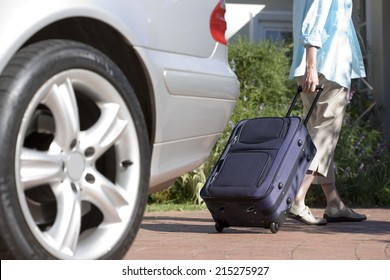 Senior Woman Pulling Suitcase On Wheels From Parked Car Boot On Driveway, Rear View (surface Level)