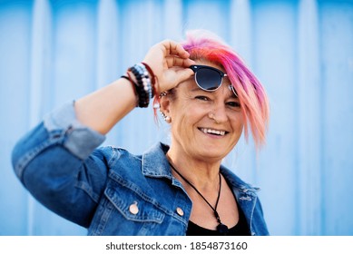 Senior Woman With Purple Pink Hair And Sunglasses Standing Against Blue Background.