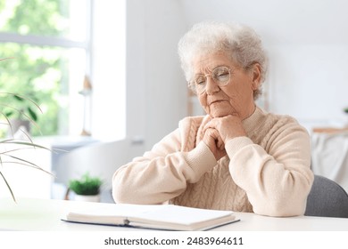 Senior woman reading Bible at table in living room - Powered by Shutterstock