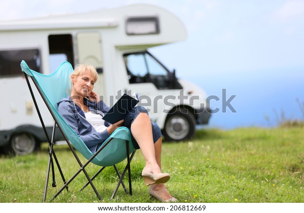 Senior woman\
reading book, camper in\
background