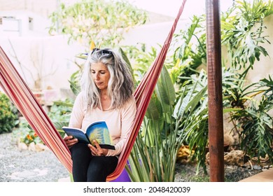 Senior woman reading book sitting on hammock at front or back yard. Elderly woman relaxing at garden. Old woman sitting on swing hammock at her house garden and reading a book
