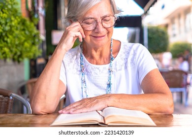 Senior woman reading a book sitting outdoor at a cafe table - smiling caucasian elderly lady in white t-shirt and glasses - Shutterstock ID 2231420139