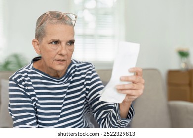 Senior Woman Reading a Receipt in Living Room with Concerned Expression, Wearing Striped Shirt and Glasses on Head - Powered by Shutterstock