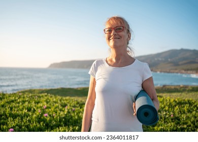 Senior woman ready for yoga exercises on seashore. Pensioner yogini holding yogamat. Mature woman going to practise yoga at sunrise at beach. Concept of healthy lifestyle on retirement - Powered by Shutterstock