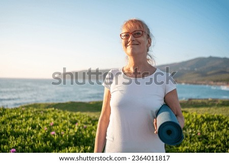 Senior woman ready for yoga exercises on seashore. Pensioner yogini holding yogamat. Mature woman going to practise yoga at sunrise at beach. Concept of healthy lifestyle on retirement