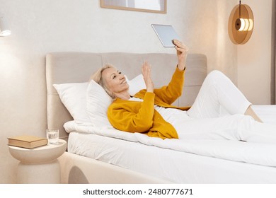 Senior Woman Relaxing In Bed, Using Tablet For Video Call And Smiling - Powered by Shutterstock