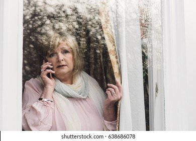 Senior Woman Is Reporting A Neighborhood Crime To The Police On The Phone. She Is Looking Out Her Window. 