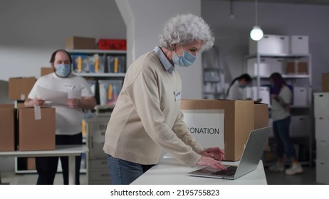 Senior Woman In Safety Mask Type On Laptop In Donation Warehouse. Volunteers In Protective Face Mask Work In Charitable Distribution Center