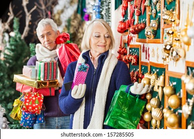 Senior Woman Shopping Christmas Ornaments At Store With Man Holding Presents In Background