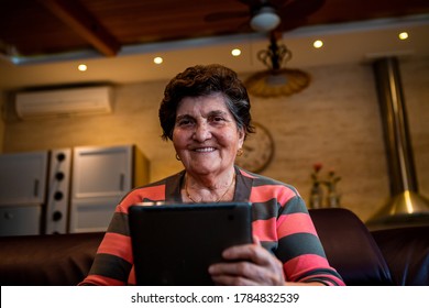 A Senior Woman With Short Black Hair Is Sitting In A Sofa In Her Home And Has A Tablet Laptop In Her Hand. She Is Very Happy To Talk To Her Family Via Video Call.