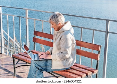 Senior woman with short gray hair, wear jeans and hoodie, sitting on bench near lake, using laptop. Working online, using some online services, online shopping. Technology, elderly, concept - Shutterstock ID 2246004321