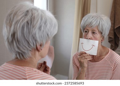 A senior woman with short gray hairstyle is holding a drawing with a smile in front of the mirror - Powered by Shutterstock