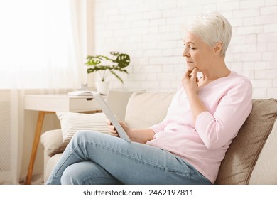 A senior woman with short white hair is seated on a beige couch, holding and using tablet with a thoughtful expression. She is casually dressed in a pink top and blue jeans - Powered by Shutterstock