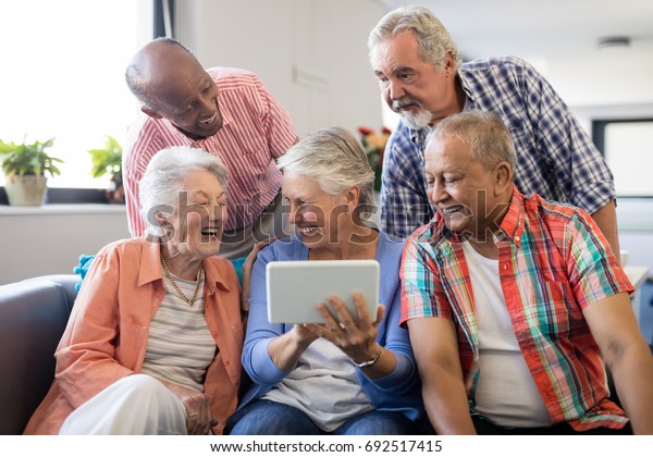 Senior woman showing digital tablet\
to cheerful friends while sitting on sofa at nursing\
home