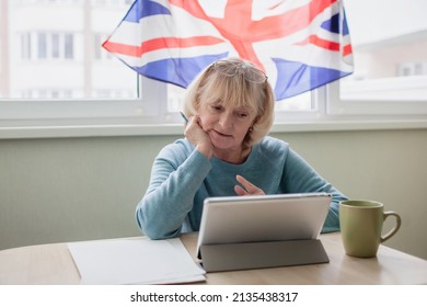 Senior Woman Sits At Desk Before Laptop And Learns English Via Internet, Language Classes For Ukrainian Refugee For Better Adaptation Abroad, British Flag On The Window, Social Distance Education