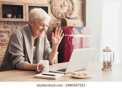 A senior woman sits at a kitchen table with a laptop in front of her. She is holding a cup of coffee in her left hand and waving with her right hand, indicating she is video chatting with someone. - Powered by Shutterstock