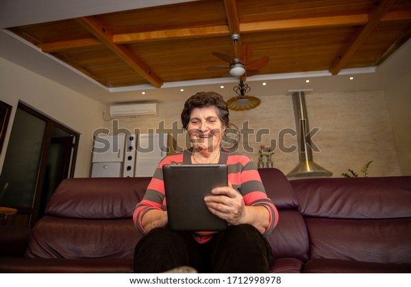 A senior woman sits\
on a couch in an apartment and holds a laptop computer tablet. She\
is cheerful and laughing because something funny is looking up on\
the internet.