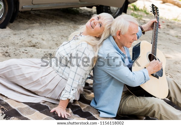 senior woman sitting back to back with man playing\
guitar on sand