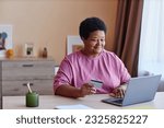 Senior woman sitting by desk in front of laptop in living room and entering data of her credit card while ordering products in online shop