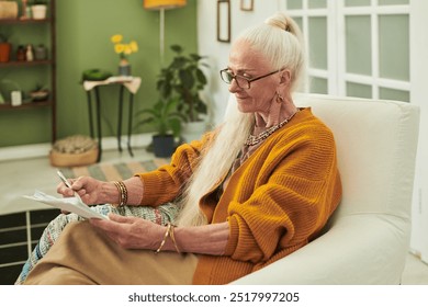 Senior woman sitting in comfortable chair writing notes with pen in hand, surrounded by cozy home decor and plants in background - Powered by Shutterstock