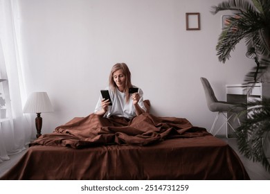 Senior woman sitting comfortably in bed holding a credit card and smartphone, making an online purchase, enjoying a relaxed morning in her cozy bedroom. - Powered by Shutterstock