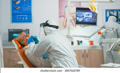 Senior Woman Sitting In Dental Clinic Taking Care Of Tooth Health During Covid-19 Pandemic. Concept Of New Normal Dentist Visit In Coronavirus Outbreak Wearing Protective Suit And Face Shield