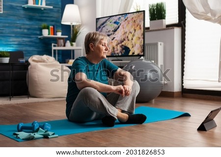 Senior woman sitting in lotus position on yoga mat training body muscles slimming weight after watching online fitness lesson using tablet computer. Pensioner stretching in living room