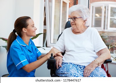 Senior Woman Sitting In Motorized Wheelchair Talking With Nurse In Retirement Home